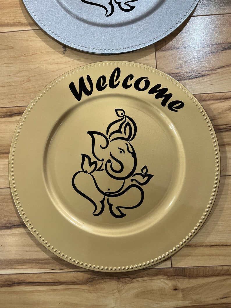 May include: Gold charger plate with a black outline of Ganesha and the word "Welcome" in black cursive lettering.