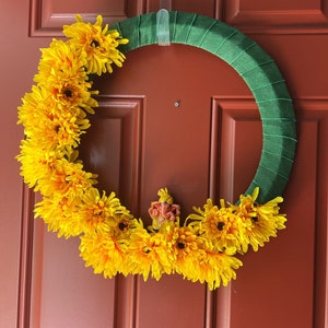 Puede incluir: Una corona de flores amarillas con una peque&ntilde;a figura naranja en el centro, colgada en una puerta de madera marr&oacute;n.