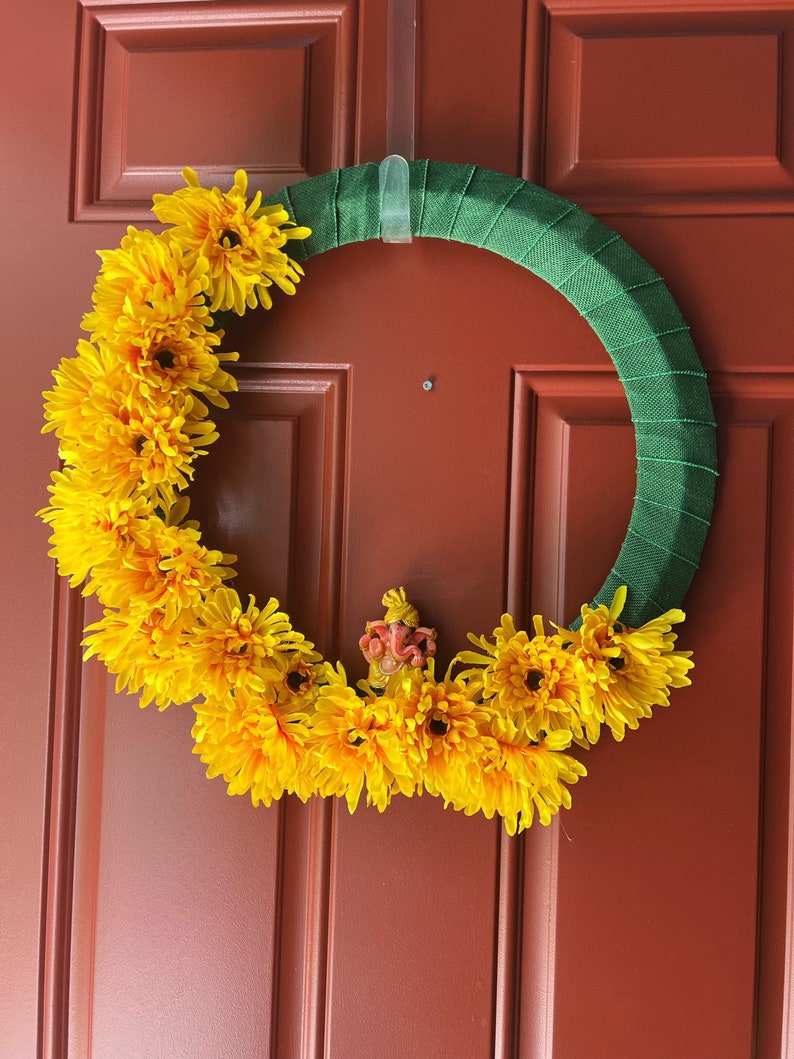 Puede incluir: Una corona de flores amarillas con una peque&ntilde;a figura naranja en el centro, colgada en una puerta de madera marr&oacute;n.
