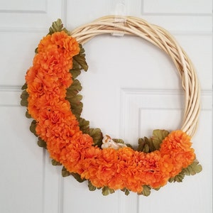 Puede incluir: Una corona hecha de ramas trenzadas con un c&iacute;rculo de flores de seda naranja y hojas verdes.