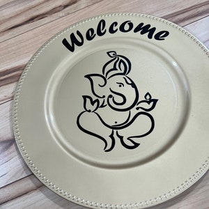May include: A gold charger plate with a black outline of the Hindu deity Ganesha and the word "Welcome" in black lettering.