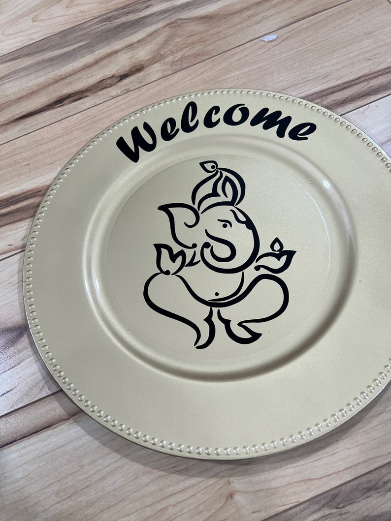 May include: A gold charger plate with a black outline of the Hindu deity Ganesha and the word "Welcome" in black lettering.