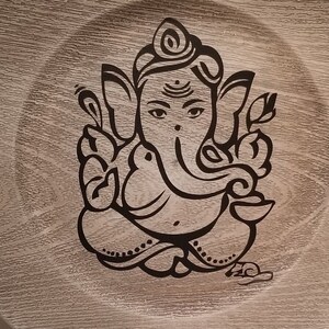 May include: A round wooden plate with a black and white image of the Hindu deity Ganesha. The plate has a wood grain pattern.