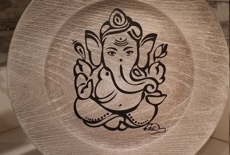 May include: A round wooden plate with a black and white image of the Hindu deity Ganesha. The plate has a wood grain pattern.