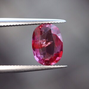 May include: A vibrant pink oval-shaped gemstone held by tweezers. The gemstone has a faceted cut and is translucent.
