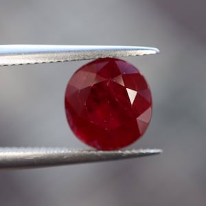 Puede incluir: Una piedra preciosa de rubí rojo oscuro sostenida en unas pinzas. El rubí tiene forma ovalada y un corte facetado.