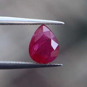 以下が含まれることがあります： ピンセットで保持された、ファセットカットされた洋梨型のルビーの宝石。ルビーは濃い赤色で、光沢のある仕上がりです。