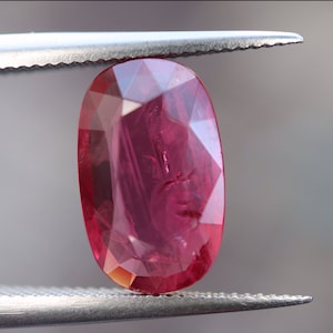 May include: A vibrant red oval-shaped gemstone with a faceted cut. The gemstone is held in place by a pair of tweezers.