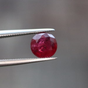 May include: A deep red ruby gemstone with a faceted cut, held in tweezers.