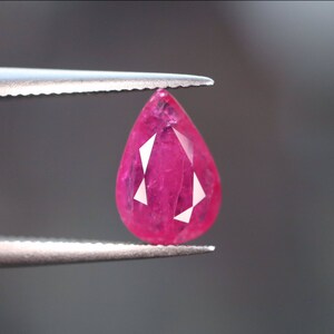 May include: A pear-shaped pink ruby gemstone held by tweezers.