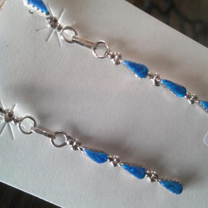 May include: A pair of silver dangle earrings with blue turquoise stones. The earrings are on a white background with a tag that says "INDIAN HAND STERLING 83-2373 ER2373".