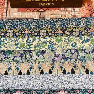 Könnte beinhalten: Ein Stapel Liberty-Stoffe mit verschiedenen floralen und geometrischen Mustern in Blautönen, Grün- und Rosatönen. Die Stoffe sind in Schichten angeordnet, wobei sich oben eine Holzkiste mit der Aufschrift "Liberty Fabrics" befindet.