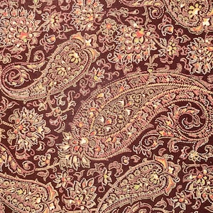 May include: Close-up of a fabric with a paisley pattern. The design features large, teardrop-shaped motifs and floral elements in shades of brown, gold, and orange against a dark brown background. The fabric has a detailed, textured appearance.