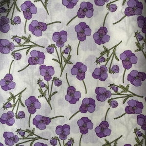 Op de afbeelding: Een stof met een herhalend bloemenpatroon. Het ontwerp toont paarse viooltjes met groene stelen en bladeren op een witte achtergrond. De viooltjes hebben een gedetailleerde bloembladstructuur.