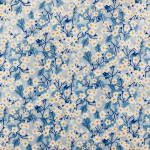 Puede incluir: Primer plano de una tela con un estampado floral. El diseño presenta pequeñas flores blancas con centros amarillos y hojas y tallos azules sobre un fondo azul claro. El patrón es denso y cubre toda la superficie.