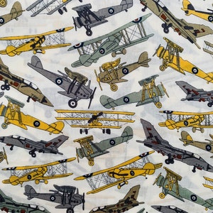 May include: A white fabric with a repeating pattern of various vintage aeroplanes in shades of yellow, green, and grey. The aeroplanes are in different sizes and orientations, creating a dynamic and whimsical design.