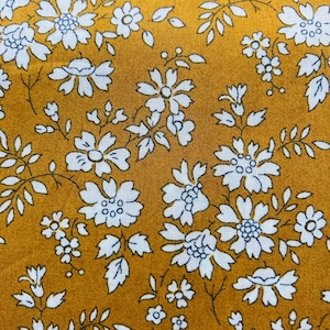May include: A mustard yellow fabric with a white floral pattern. The fabric is covered in small white flowers with blue centers.