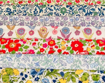 May include: A stack of Liberty Fabrics featuring various floral patterns in shades of blue, red, yellow, and green. The fabrics are arranged in a neat stack with a wooden box labeled "Liberty Fabrics" on top.