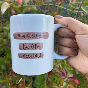 Op de afbeelding: Witte keramische mok met een ontwerp van bruine en roze penseelstreken. De mok heeft de tekst "manifesting the life i deserve!" erop gedrukt.