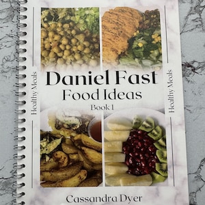 Può includere: Un libro rilegato a spirale intitolato "Daniel Fast Food Ideas Book 1" con immagini di pasti sani. La copertina del libro presenta uno sfondo bianco con un motivo a marmo e il testo "Healthy Meals" sui lati sinistro e destro. Il libro è di Cassandra Dyer e ha l'account social @cassisscreative.