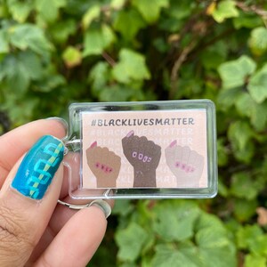 May include: A clear plastic keychain with a beige background and three raised fists in different skin tones. The fists have pink fingernails and the text "#BlackLivesMatter" is printed in black at the top.