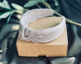 May include: A light gray linen knotted headband on a brown cardboard box. The headband is made of a soft, woven fabric and has a simple, elegant design.