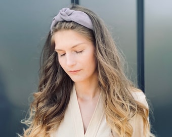 May include: A woman wearing a light brown linen dress with a knotted headband in a dusty purple color. The dress has a V-neckline, long sleeves, and a belt around the waist.