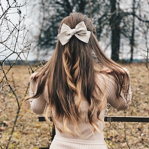 Könnte beinhalten: Eine Frau mit langen braunen Haaren trägt ein beigefarbenes Strickkleid mit einer großen beigefarbenen Schleife im Haar.