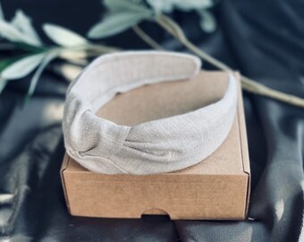 May include: A beige linen headband with a knotted detail, resting on a small brown cardboard box. The headband is a neutral color, and the box is plain. The background is a dark, textured fabric with a sprig of greenery.