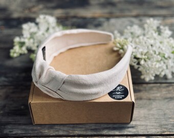 May include: A light beige knotted headband, likely linen, sits atop a brown cardboard box. The headband has a simple, elegant design. The box has a black and white logo that says "ACCESSORIES & MORE". White flowers are in the background.
