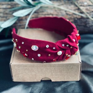 Op de afbeelding: Een bordeauxrode hoofdband met een geknoopt ontwerp, versierd met strass, parels en rode edelstenen.