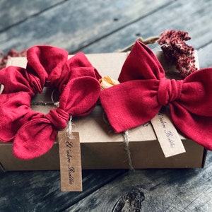 Puede incluir: Dos scrunchies de pelo de tela roja y un lazo de tela roja, empaquetados en una caja marrón con una etiqueta "L'adore - Ruffles".