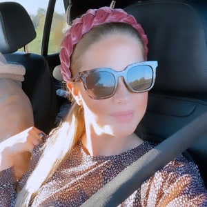Peut inclure: Une femme portant un bandeau en velours rose et des lunettes de soleil en &eacute;caille de tortue dans une voiture. Elle porte une chemise &agrave; motifs marrons et blancs et une ceinture de s&eacute;curit&eacute; noire.