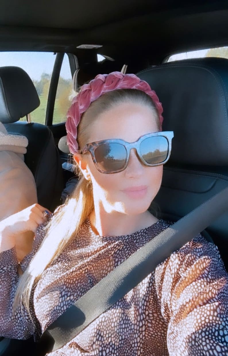 Peut inclure: Une femme portant un bandeau en velours rose et des lunettes de soleil en &eacute;caille de tortue dans une voiture. Elle porte une chemise &agrave; motifs marrons et blancs et une ceinture de s&eacute;curit&eacute; noire.