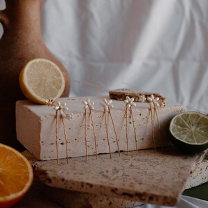 Puede incluir: Un juego de seis horquillas doradas con detalles de flores de perlas. Las horquillas est&aacute;n dispuestas sobre una superficie de piedra marr&oacute;n claro con un lim&oacute;n y una lima en el fondo.