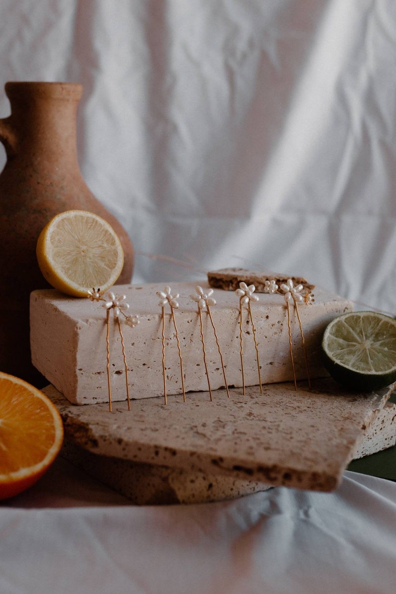 Puede incluir: Un juego de seis horquillas doradas con detalles de flores de perlas. Las horquillas est&aacute;n dispuestas sobre una superficie de piedra marr&oacute;n claro con un lim&oacute;n y una lima en el fondo.