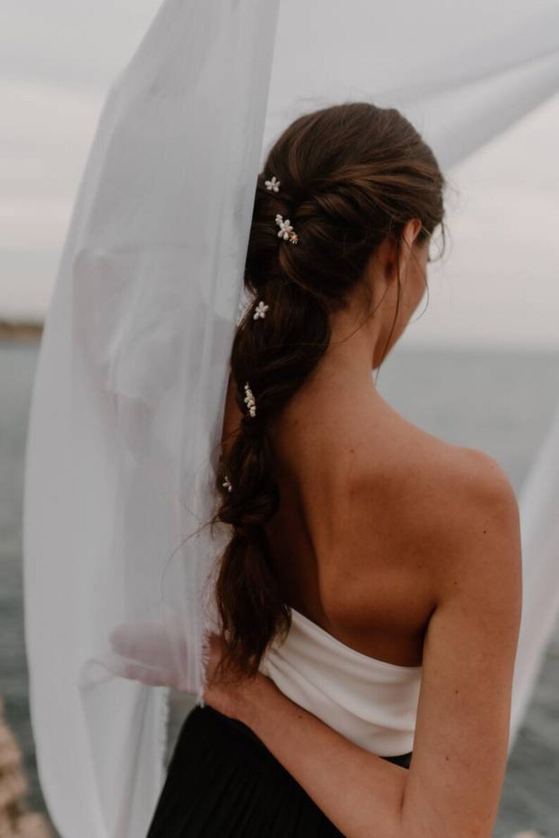 Puede incluir: Una mujer con el pelo largo y casta&ntilde;o peinado en una trenza con peque&ntilde;as pinzas para el pelo con forma de flores blancas. Lleva un top blanco sin tirantes y una falda negra.
