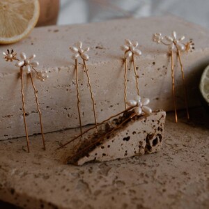 Puede incluir: Conjunto de seis horquillas doradas con detalles de flores de perlas. Las horquillas est&aacute;n dispuestas sobre una superficie texturizada.