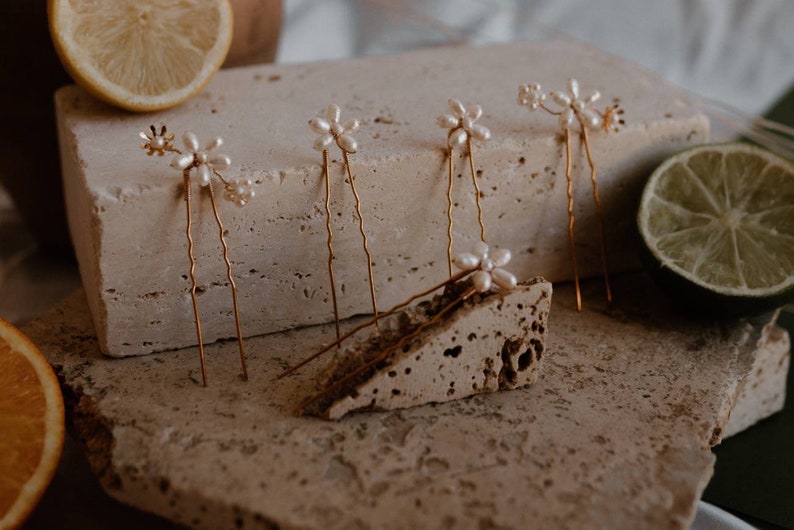 Puede incluir: Conjunto de seis horquillas doradas con detalles de flores de perlas. Las horquillas est&aacute;n dispuestas sobre una superficie texturizada.