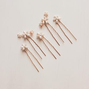 Puede incluir: Conjunto de ocho horquillas doradas con detalles de flores de perlas y oro. Las horquillas est&aacute;n dispuestas en forma de abanico sobre un fondo blanco.