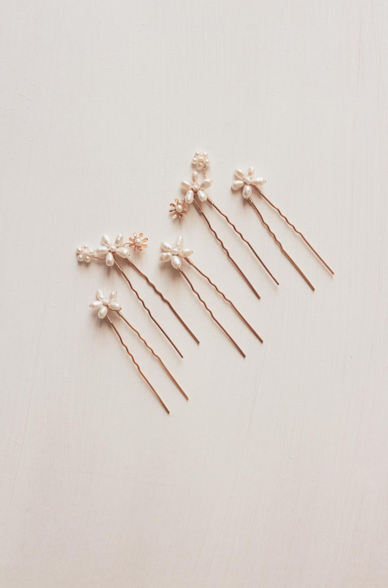 Puede incluir: Conjunto de ocho horquillas doradas con detalles de flores de perlas y oro. Las horquillas est&aacute;n dispuestas en forma de abanico sobre un fondo blanco.