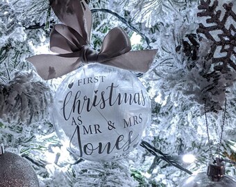 Puede incluir: Un adorno de Navidad de vidrio transparente con una cinta blanca y el texto "First Christmas as Mr & Mrs Jones" en un árbol de Navidad nevado.