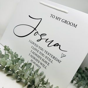 May include: White gift bag with a black handwritten message that says "To My Groom Joshua I Loved You Yesterday I Love You Still I Always Have I Always Will 27.10.2023" with a small heart symbol.