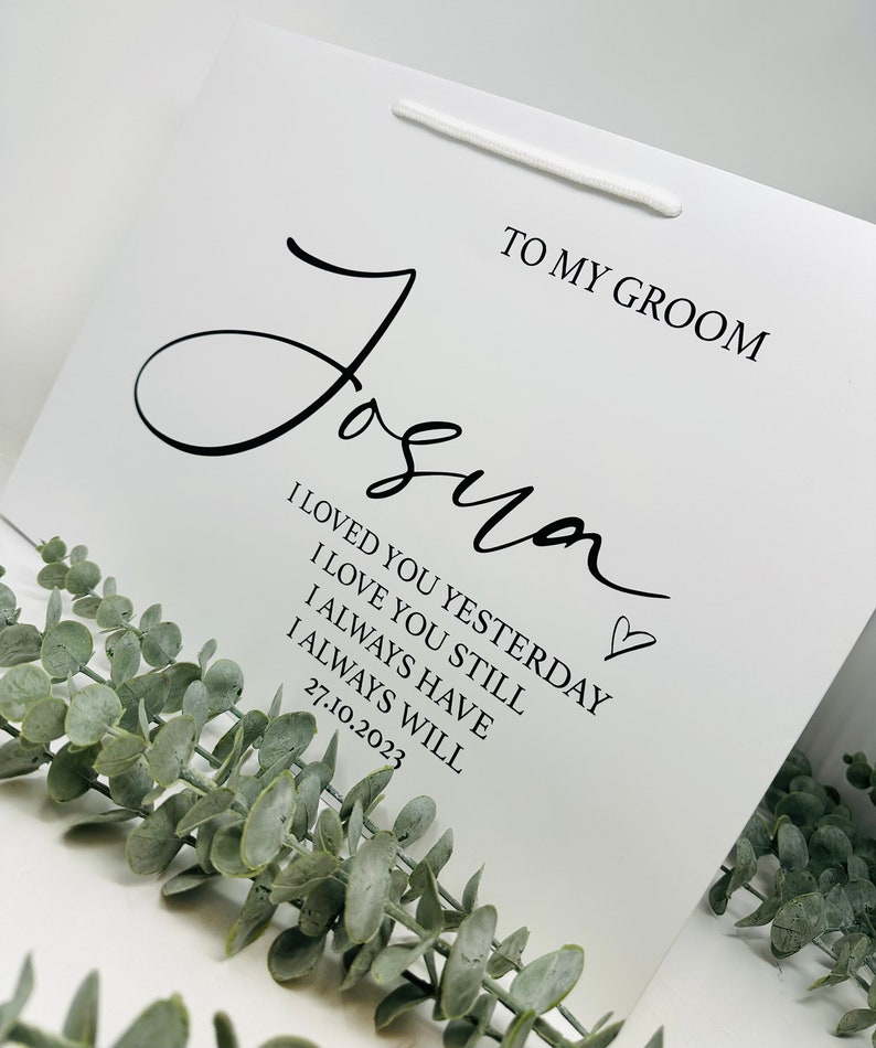 May include: White gift bag with a black handwritten message that says "To My Groom Joshua I Loved You Yesterday I Love You Still I Always Have I Always Will 27.10.2023" with a small heart symbol.