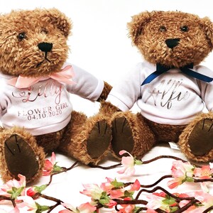Puede incluir: Dos osos de peluche marrones que llevan camisas blancas con nombres personalizados y el texto "Flower Girl" y "04.10.2020". Los osos están sentados sobre una superficie blanca con flores rosas.