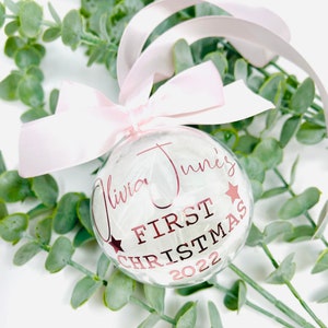 Puede incluir: Un adorno de Navidad de vidrio transparente con plumas blancas en su interior. El adorno est&aacute; decorado con una cinta rosa y letras doradas que dicen "Olivia June's First Christmas 2022".