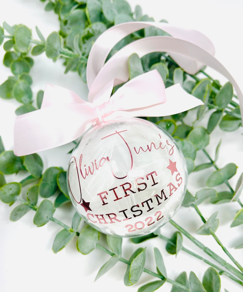 Puede incluir: Un adorno de Navidad de vidrio transparente con plumas blancas en su interior. El adorno est&aacute; decorado con una cinta rosa y letras doradas que dicen "Olivia June's First Christmas 2022".