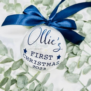 Puede incluir: Un adorno de Navidad de vidrio transparente con fondo blanco y letras azules. El adorno dice "Ollie's First Christmas 2023" y tiene dos estrellas azules a cada lado del texto.