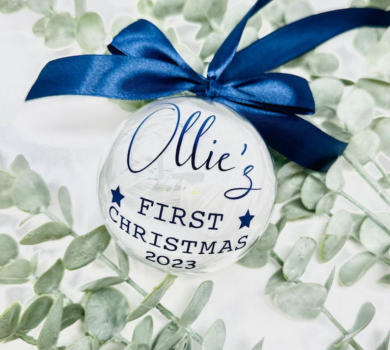 Puede incluir: Un adorno de Navidad de vidrio transparente con fondo blanco y letras azules. El adorno dice "Ollie's First Christmas 2023" y tiene dos estrellas azules a cada lado del texto.
