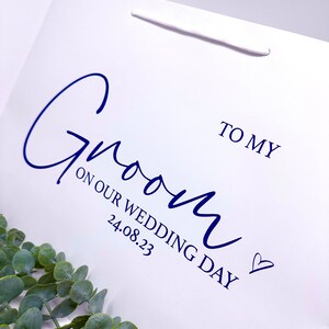 Puede incluir: Una bolsa de regalo blanca con una inscripci&oacute;n azul que dice "To my Groom on our Wedding Day 24.08.23" con un peque&ntilde;o s&iacute;mbolo de coraz&oacute;n.